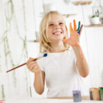 happy-playful-cute-freckled-blonde-girl-dressed-white-holding-brush-one-hand-showing-another-hand-which-she-messed-up-with-paint